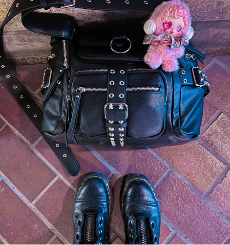 Black studded handbag with a pink charm beside black boots on a brick floor.